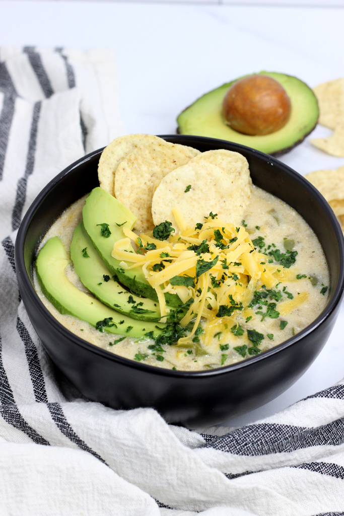 A large bowl of enchilada soup, with toppings. Sliced avocado, shredded cheese, tortilla chips and cilantro.